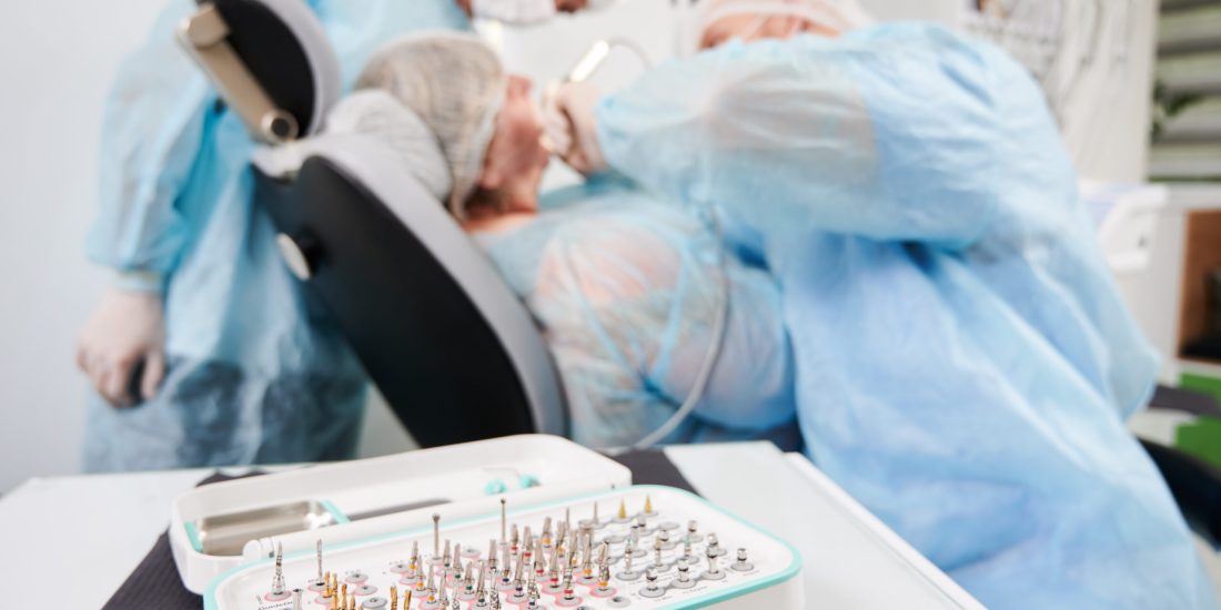 Horizontal close-up snapshot of a surgical taper kit, opened on dentist table, in focus. Dentist and his assistant during surgery performance on blurred background. Concept of oral care