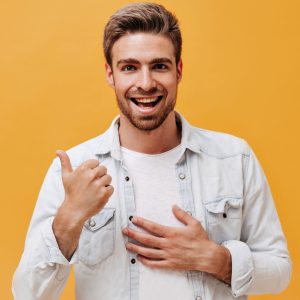 Cheerful guy with beard in white t-shirt and denim jacket looking into camera and smiling on orange isolated background..